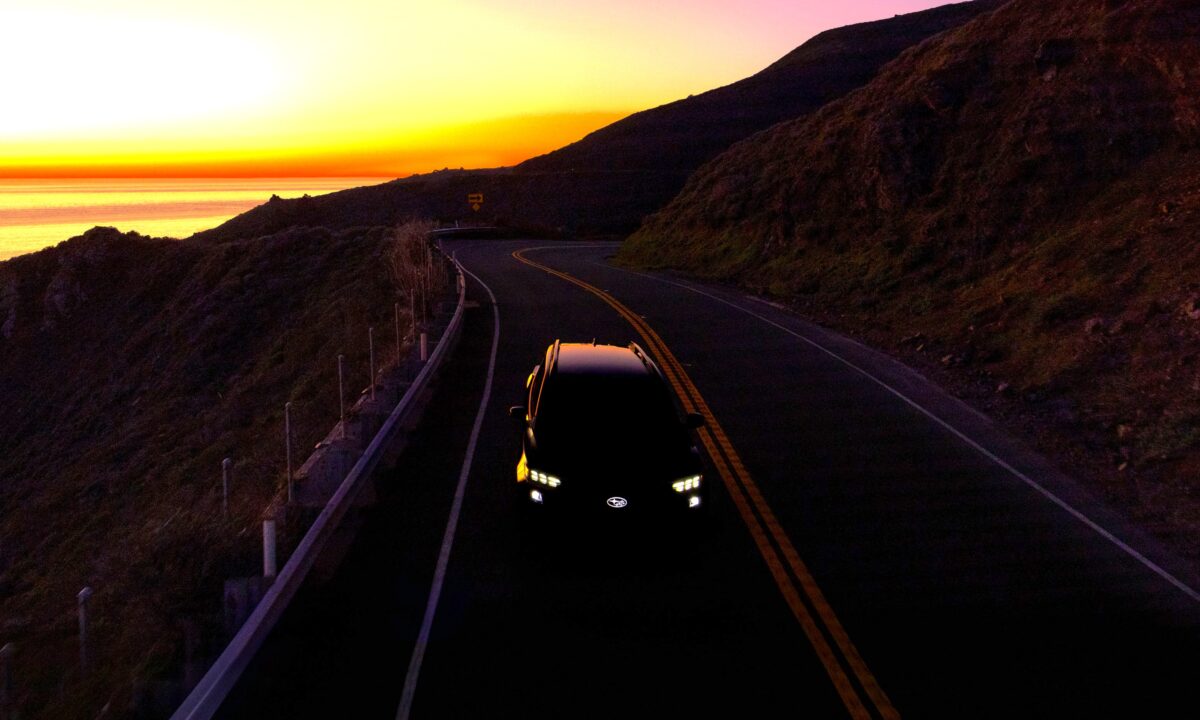 רכב פנאי של סובארו מצולם מלמעלה בכביש הררי מתפתל בשעת שקיעה, כאשר חזית הרכב מוארת בפנסי לד והים והאופק נראים ברקע
