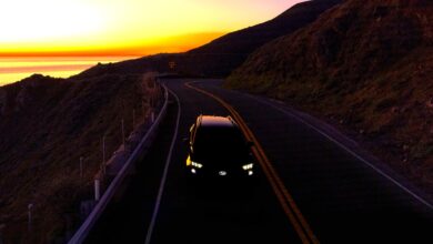 רכב פנאי של סובארו מצולם מלמעלה בכביש הררי מתפתל בשעת שקיעה, כאשר חזית הרכב מוארת בפנסי לד והים והאופק נראים ברקע